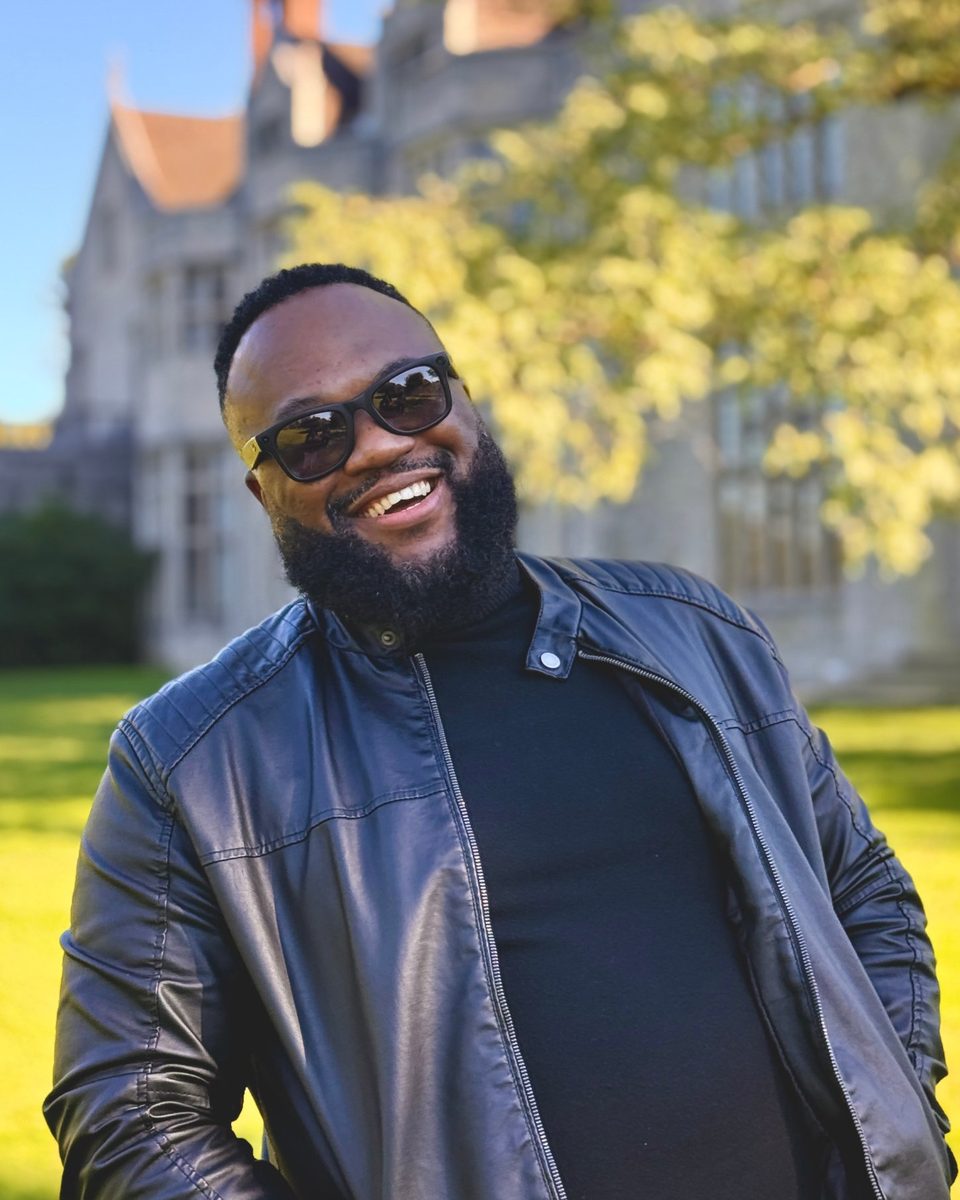 Shane S. Scott smiling in sunglasses and a leather jacket outside a historic stone manor